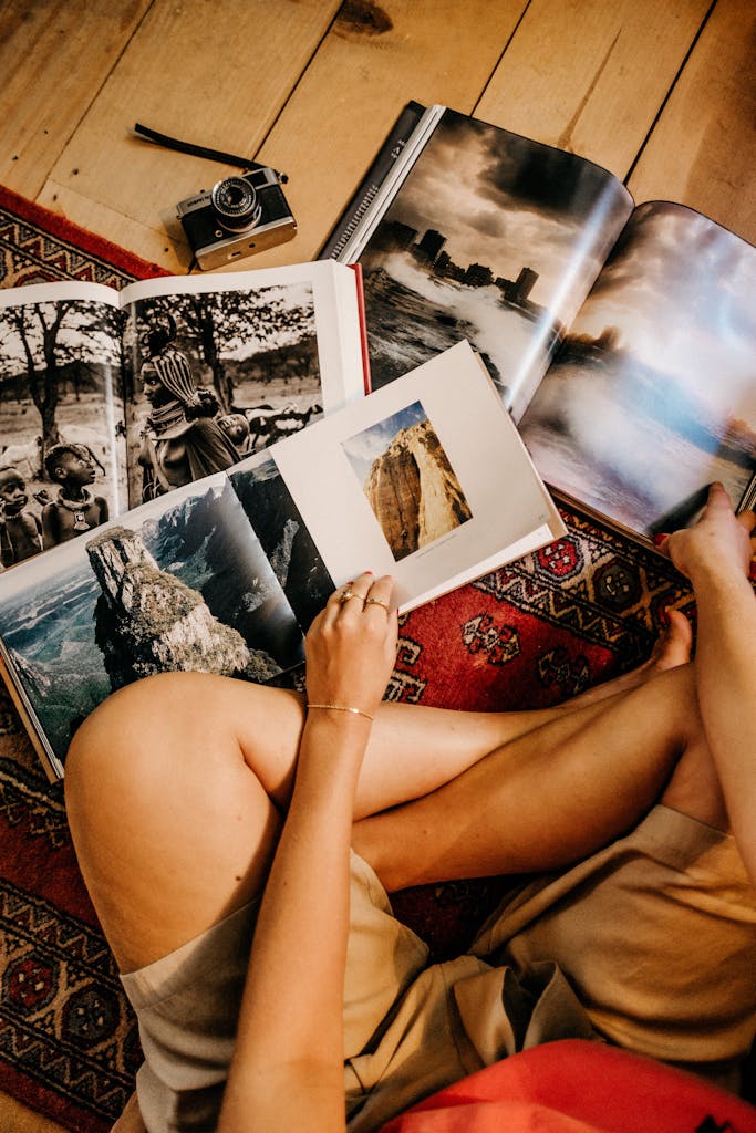 Person sitting on a rug, browsing photo books on a wooden floor with vintage camera nearby.